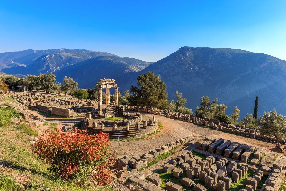 Ancient ruins of Delphi with a circular temple structure, set against a backdrop of mountains and clear blue sky.